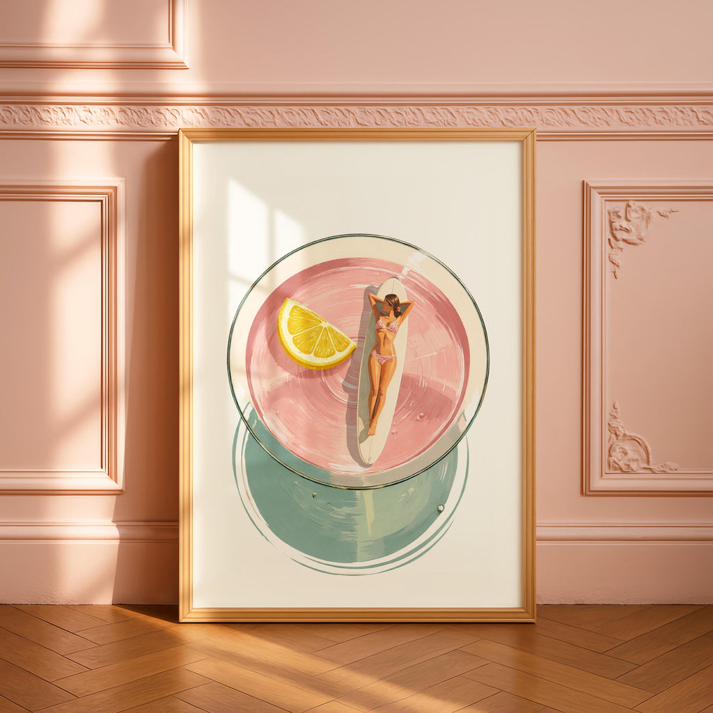 May include: A framed art print depicts a cocktail with a woman sunbathing on a surfboard inside. The drink is pink with a lemon wedge. The bottom of the glass is a teal circle. The frame is gold.