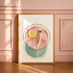 May include: A framed art print depicts a cocktail with a woman sunbathing on a surfboard inside. The drink is pink with a lemon wedge. The bottom of the glass is a teal circle. The frame is gold.